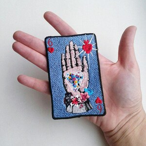 May include: A blue denim patch with a hand holding a heart with a red outline. The hand is surrounded by red hearts and the number 6 is in the top left corner. The patch is embroidered with colorful details.