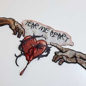 May include: Two embroidered hands reaching towards each other, with a heart pierced by barbed wire and a banner reading "TEAR ME APART" in between.