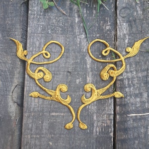 Könnte beinhalten: Zwei goldfarbene, verzierte, gestickte Applikationen in Form eines stilisierten Blumenmusters. Die Applikationen befinden sich auf einer Holzoberfläche.