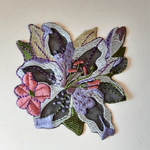 Pode incluir: Uma delicada aplicação de flores bordadas que apresenta um grupo de lírios em tons de roxo, rosa e verde. A aplicação é feita de tecido transparente e tem um design detalhado.