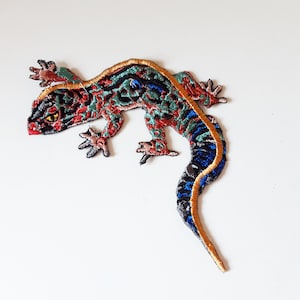 May include: A colourful embroidered gecko patch with a gold outline. The gecko is in a curled position with its tail wrapped around its body. The patch is ideal for adding a touch of whimsy to clothing, bags, or other accessories.