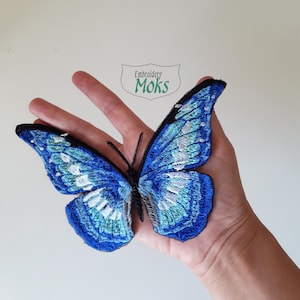 Puede incluir: Parche de mariposa bordada en tonos azules, blancos y negros. La mariposa tiene patrones de alas detallados y un cuerpo negro. El parche mide aproximadamente 15 cm de ancho. El texto "Embroidery Moks" está en la esquina superior izquierda.