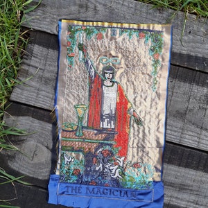 May include: Embroidered tarot card depicting The Magician. The card features a man in a red robe holding a wand, with a table in front of him and a cup, a sword, a pentagram, and a coin on the table. The card is surrounded by a gold border and a blue background.