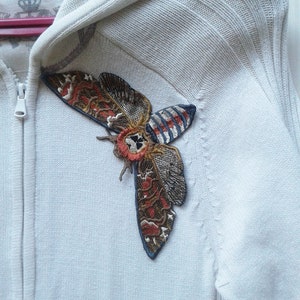 Può includere: Una felpa con cappuccio bianca con un grande disegno di falena ricamato sul lato sinistro. La falena è marrone, arancione e blu con un motivo a forma di teschio sulle ali.