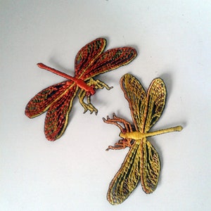 May include: Two embroidered dragonfly patches. One dragonfly is primarily red and yellow, the other is yellow and black. The wings have a detailed, openwork design. The dragonflies are on a white background.