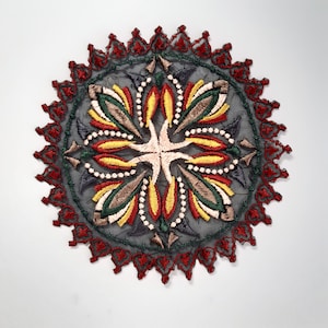 Peut inclure: Un patch brodé circulaire avec un motif floral dans des tons de rouge, jaune, vert et marron. Le centre du motif est une forme de croix avec un fond blanc. Le patch est entouré d'une bordure rouge avec un bord festonné.