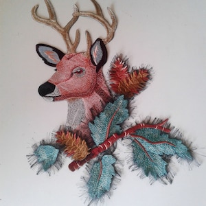 Op de afbeelding: Geborduurde strijk-patch met een roze hertenkop met geweien, omringd door groene en bruine bladeren.
