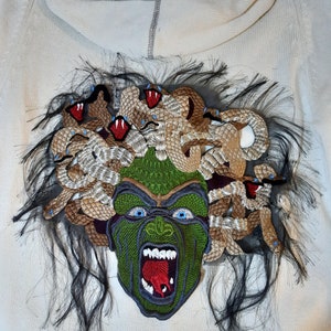 May include: A white hoodie with a green and brown embroidered patch of Medusa's head with snakes for hair. The patch has a detailed design with red and blue accents.