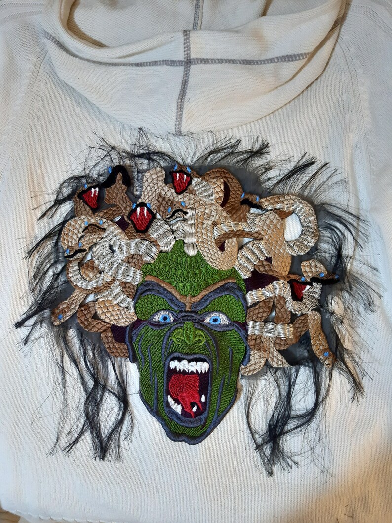 May include: A white hoodie with a green and brown embroidered patch of Medusa's head with snakes for hair. The patch has a detailed design with red and blue accents.