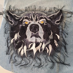 May include: Embroidered bear patch in black, grey, and white, with yellow eyes, on a light blue denim background. The patch is framed with black fringe, creating a textured effect.