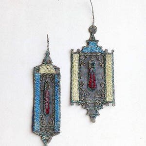 K&ouml;nnte beinhalten: Zwei handgefertigte Laternen-Ornamente. Die Laternen weisen aufwendige silberfarbene Metallarbeiten mit blauen, gelben und roten Akzenten auf. Das Design beinhaltet ein quastenartiges Element in der Mitte. Der Hintergrund ist wei&szlig;.