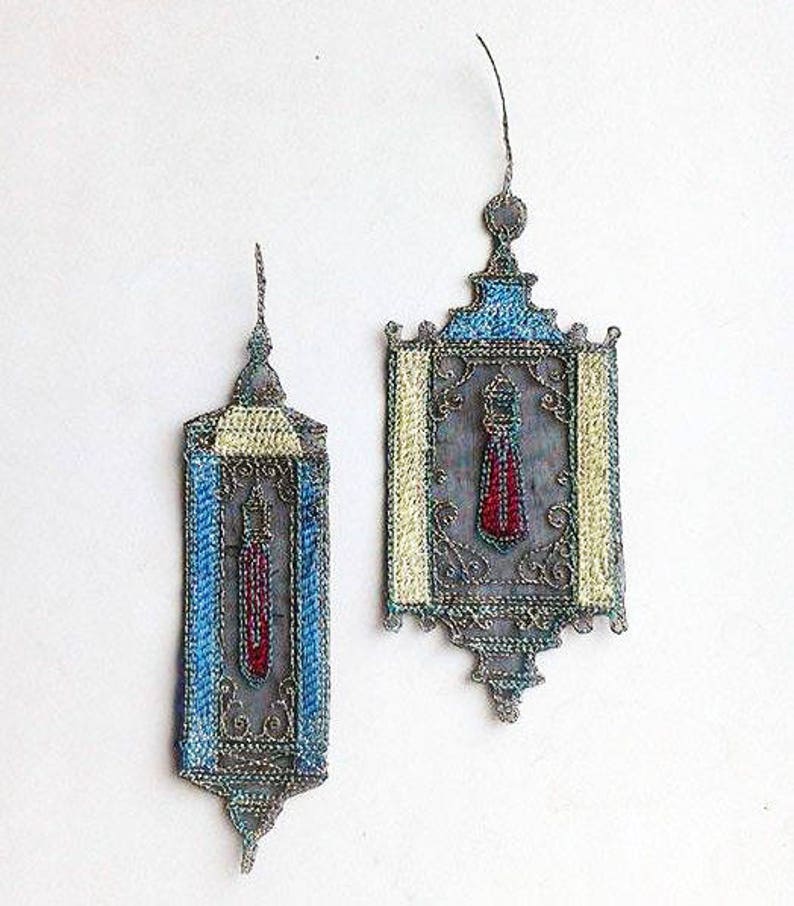 K&ouml;nnte beinhalten: Zwei handgefertigte Laternen-Ornamente. Die Laternen weisen aufwendige silberfarbene Metallarbeiten mit blauen, gelben und roten Akzenten auf. Das Design beinhaltet ein quastenartiges Element in der Mitte. Der Hintergrund ist wei&szlig;.