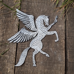 Peut inclure: Un écusson brodé argenté représentant un cheval ailé, ou pégase, avec des détails complexes. Le patch est idéal pour ajouter une touche mythique aux vêtements, sacs ou autres accessoires.