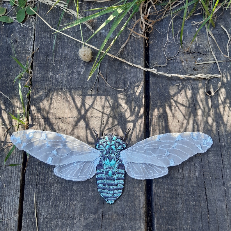 Pu&ograve; includere: Una toppa ricamata a forma di cicala con ali in pizzo azzurro chiaro e un corpo turchese e blu. L'insetto &egrave; appoggiato su una superficie di legno stagionato con erba e luce solare.