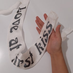 Puede incluir: Sash de tela blanca con bordado negro que dice "primer beso" y "todo sobre el amor".
