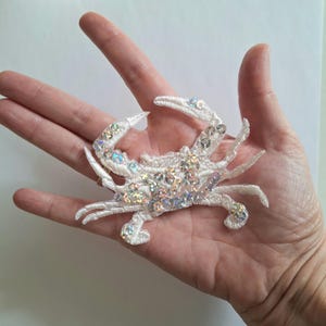 May include: White crab applique with iridescent sequins. The crab features detailed embroidery and is made of a soft, textured fabric. The crab's claws and legs are delicately shaped. Suitable for clothing or craft projects.