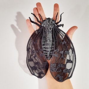 May include: Black lace cicada applique with detailed embroidery. The wings are delicate and intricate, with a black and silver color scheme.