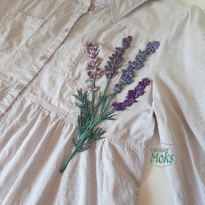 以下が含まれることがあります： ストライプの薄いグレーのシャツに、ラベンダーの花の刺繍パッチが付いている。パッチには、紫、ピンク、緑の花と緑の茎が描かれている。