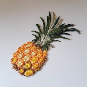 May include: A yellow and orange embroidered pineapple with green leaves. The pineapple is a decorative patch.