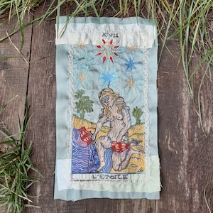 Może przedstawiać: Winietkowa, haftowana karta tarota przedstawiająca kartę Gwiazdy, z nagą kobietą wlewającą wodę z dwóch dzbanów, z gwiazdą nad głową i niebieskim tłem nieba. Karta jest oznaczona jako "XVII L'ETOILE" po francusku.