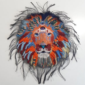 Puede incluir: Una cabeza de león bordada de colores con un tercer ojo en el centro de su frente. La melena del león está hecha de hilos de varios colores, incluyendo rojo, naranja, amarillo, azul y verde. El fondo es negro.