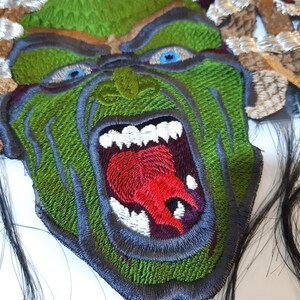 May include: Green embroidered patch of a monster with a large open mouth, showing sharp teeth and a red tongue. The monster has a green face with black eyes and is surrounded by brown and white embroidered hair.