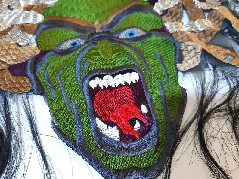 May include: Green embroidered patch of a monster with a large open mouth, showing sharp teeth and a red tongue. The monster has a green face with black eyes and is surrounded by brown and white embroidered hair.
