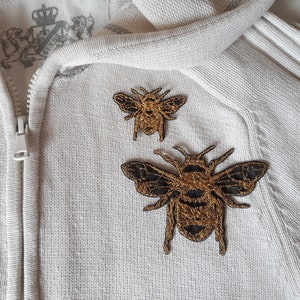 May include: Two gold and black embroidered bee patches are attached to a light beige zip-up hoodie. The bees feature detailed wing patterns and body segments. The hoodie has a Guess logo on the inside.