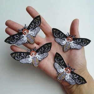 May include: Four embroidered iron-on patches of black, white, and orange moths with detailed wings and a skull-like face.