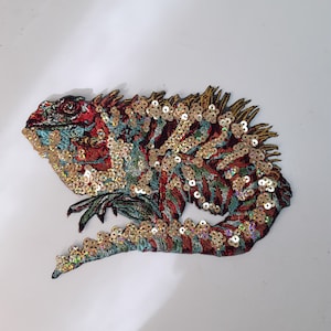 May include: A colourful, sequined iguana applique patch with red, gold, and blue accents. The iguana is sitting on a branch with its tail curled around it.