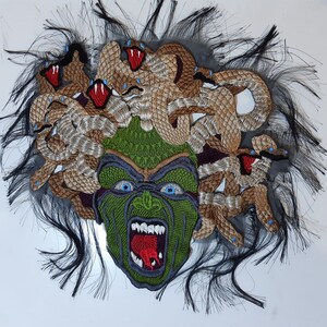 May include: A green and brown embroidered patch depicting Medusa, a mythical Greek figure with snakes for hair. The patch features a detailed face with a fierce expression and a red tongue sticking out.
