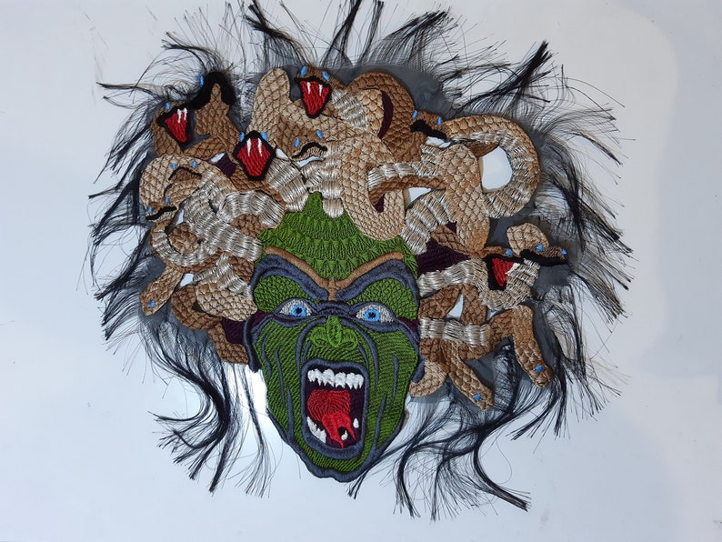 May include: A green and brown embroidered patch depicting Medusa, a mythical Greek figure with snakes for hair. The patch features a detailed face with a fierce expression and a red tongue sticking out.