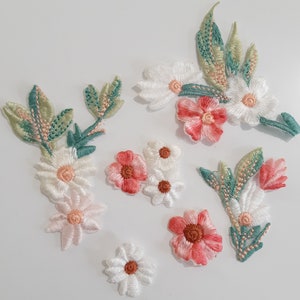 May include: Assortment of embroidered fabric flower appliques in white, pink, and green. The appliques feature various flower shapes and sizes, including daisies and roses.