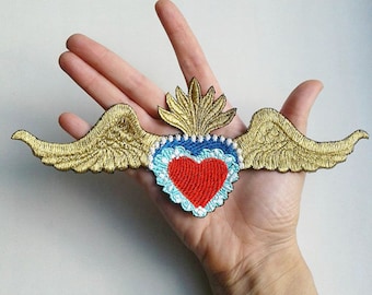 Pode incluir: Patch bordado com um coração vermelho, borda azul, asas douradas e design de chama dourada. O patch é segurado em uma mão, mostrando os detalhes do bordado. Ideal para roupas ou acessórios, este patch adiciona um toque único.