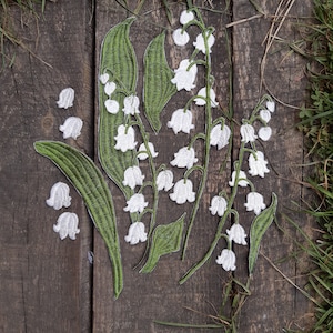 May include: A close-up of a set of white lily of the valley flower embroidery patches. The patches are arranged in a bouquet shape with green stems and leaves.