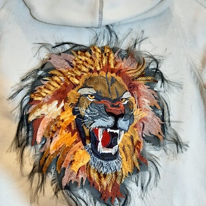 Peut inclure: Un sweat à capuche blanc avec un grand lion brodé sur le dos. Le lion rugit la gueule ouverte et a une expression féroce. Le lion est entouré d'une bordure en fourrure noire.