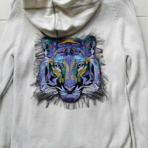 Puede incluir: Sudadera blanca con capucha con un bordado de cara de tigre de colores en la espalda.