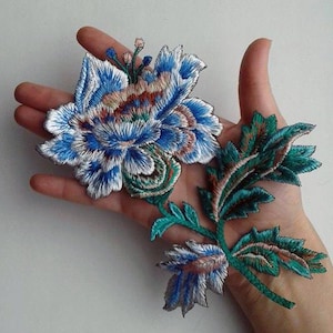 May include: A blue and green embroidered flower applique with a white center. The flower has a layered design with a variety of shades of blue and green. The applique is perfect for adding a touch of elegance to any project.