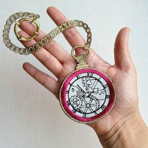 Puede incluir: Una cadena de oro con un reloj de bolsillo bordado en rosa y oro con una esfera de reloj y engranajes en su interior.