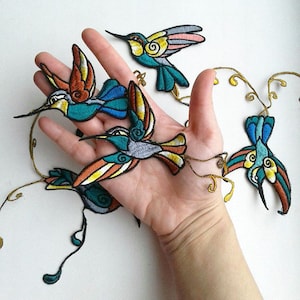 Puede incluir: Cuatro parches bordados de colibrí coloridos con detalles de hilo dorado. Los parches están dispuestos en línea en una mano.