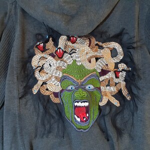 May include: A dark gray hooded sweatshirt with a large embroidered image of Medusa on the back. The embroidery features a green face with snakes for hair, red eyes, and a red tongue.