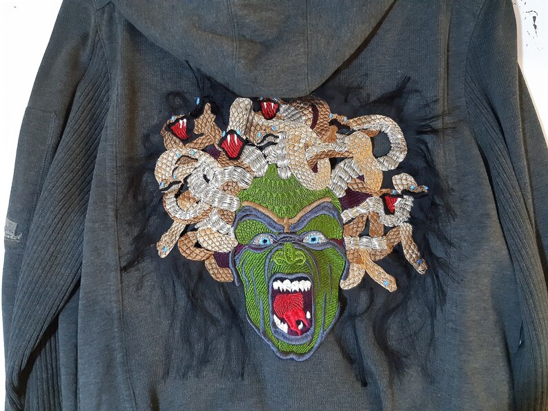 May include: A dark gray hooded sweatshirt with a large embroidered image of Medusa on the back. The embroidery features a green face with snakes for hair, red eyes, and a red tongue.