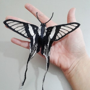 May include: A black and white butterfly with long tails, embroidered with white thread on a black fabric. The butterfly is held in a hand.