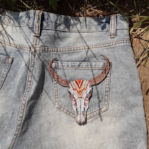 May include: A pair of blue denim jeans with a brown and white embroidered bull skull patch on the back pocket. The patch has geometric patterns in orange, red, and white.