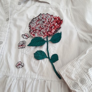 Op de afbeelding: Een wit shirt met een roze en groen geborduurd bloemenontwerp. De bloem is een hortensia met een grote bloei en kleinere bloeiwijzen op de stengel.