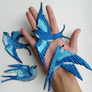 Może przedstawiać: Cztery niebieskie haftowane łatki z ptakami z białymi akcentami. Łatki mają kształt ptaków z rozłożonymi skrzydłami. Łatki znajdują się na dłoni.