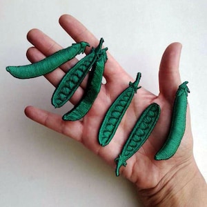 Könnte beinhalten: Sechs grüne Filz-Erbsenhülsen-Patches. Die Patches haben die Form von Erbsenhülsen und sind mit einem gestickten Detail versehen. Die Patches befinden sich auf einer Hand.
