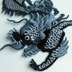 Puede incluir: Una aplicación bordada de dragón azul y negro con detalles intrincados. El dragón está enroscado con sus alas extendidas. La aplicación está hecha de una tela delicada y sería perfecta para agregar un toque de fantasía a un proyecto artesanal.