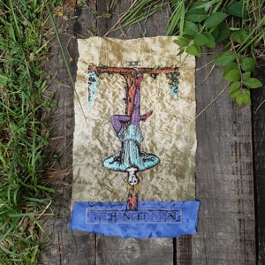 Puede incluir: Una carta de tarot vintage que representa al Ahorcado, una figura colgada boca abajo de una rama de árbol. La carta está bordada con hilos de colores sobre un fondo de tela beige. La carta está etiquetada como "El Ahorcado" en hilo azul.