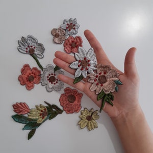 Könnte beinhalten: Sortiment an gestickten Blumen-Patches in verschiedenen Farben, darunter Weiß, Rosa, Grün und Braun. Die Patches sind auf einer weißen Oberfläche angeordnet und eine Hand hält einige der Patches.